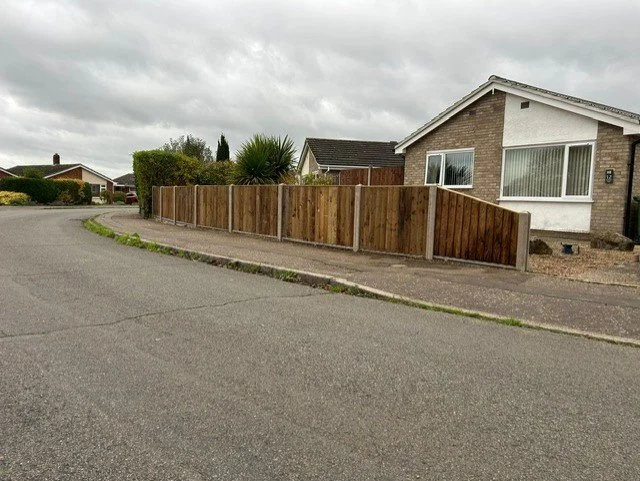 A residential street with a house and a newly installed wooden fence along the sidewalk, overcast sky.