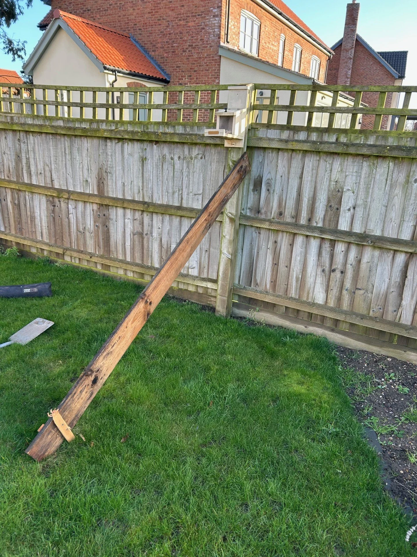 A wooden fence with a leaning fence post in a backyard with grass and houses in the background.