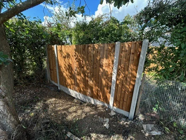 A newly installed wooden fence with concrete posts in a backyard, surrounded by trees and bushes, under a partly cloudy sky.