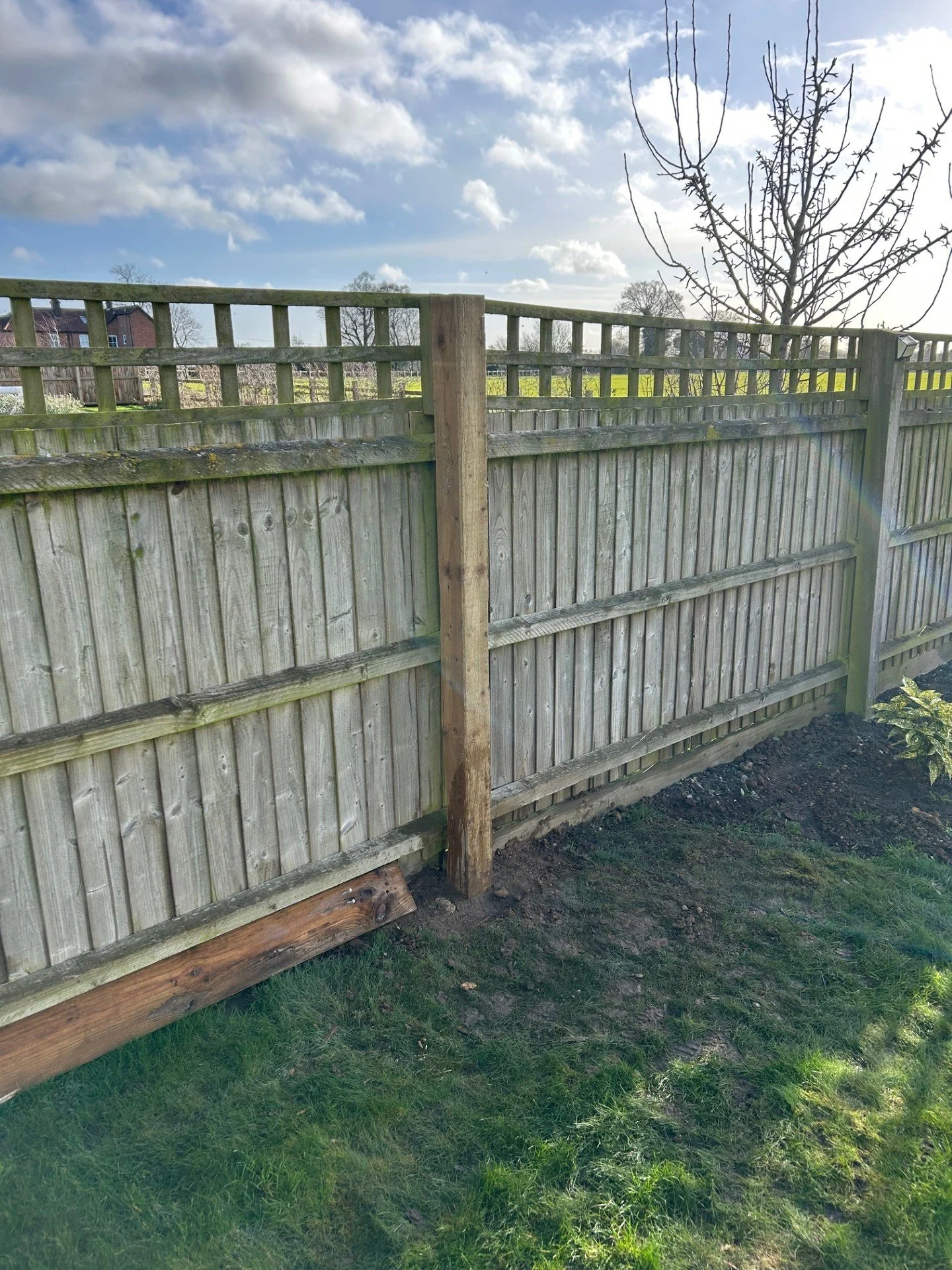 Wooden garden fence with metal rails on top, some grass and dirt in the foreground, blue sky with clouds, and a tree without leaves in the background.
