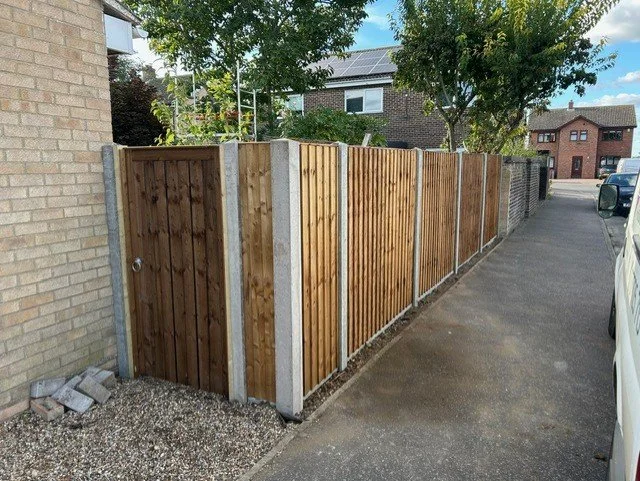 A wooden privacy fence with vertical slats runs along a sidewalk next to residential buildings and parked cars, with trees and a blue sky in the background.