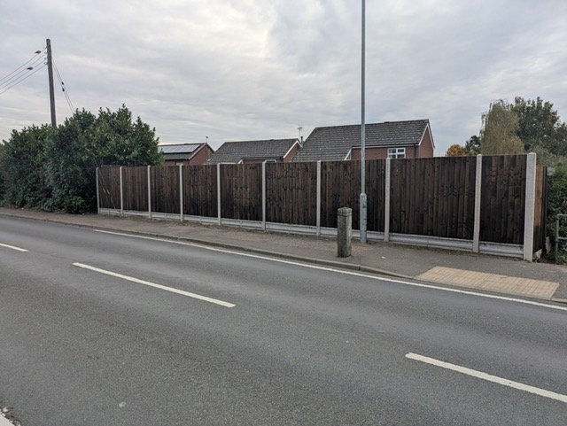 A sidewalk with a tall dark wooden fence running parallel beside a two-lane road. There is a lamppost and a small concrete post near the fence. Houses with sloped roofs are visible behind the fence. The sky is overcast.