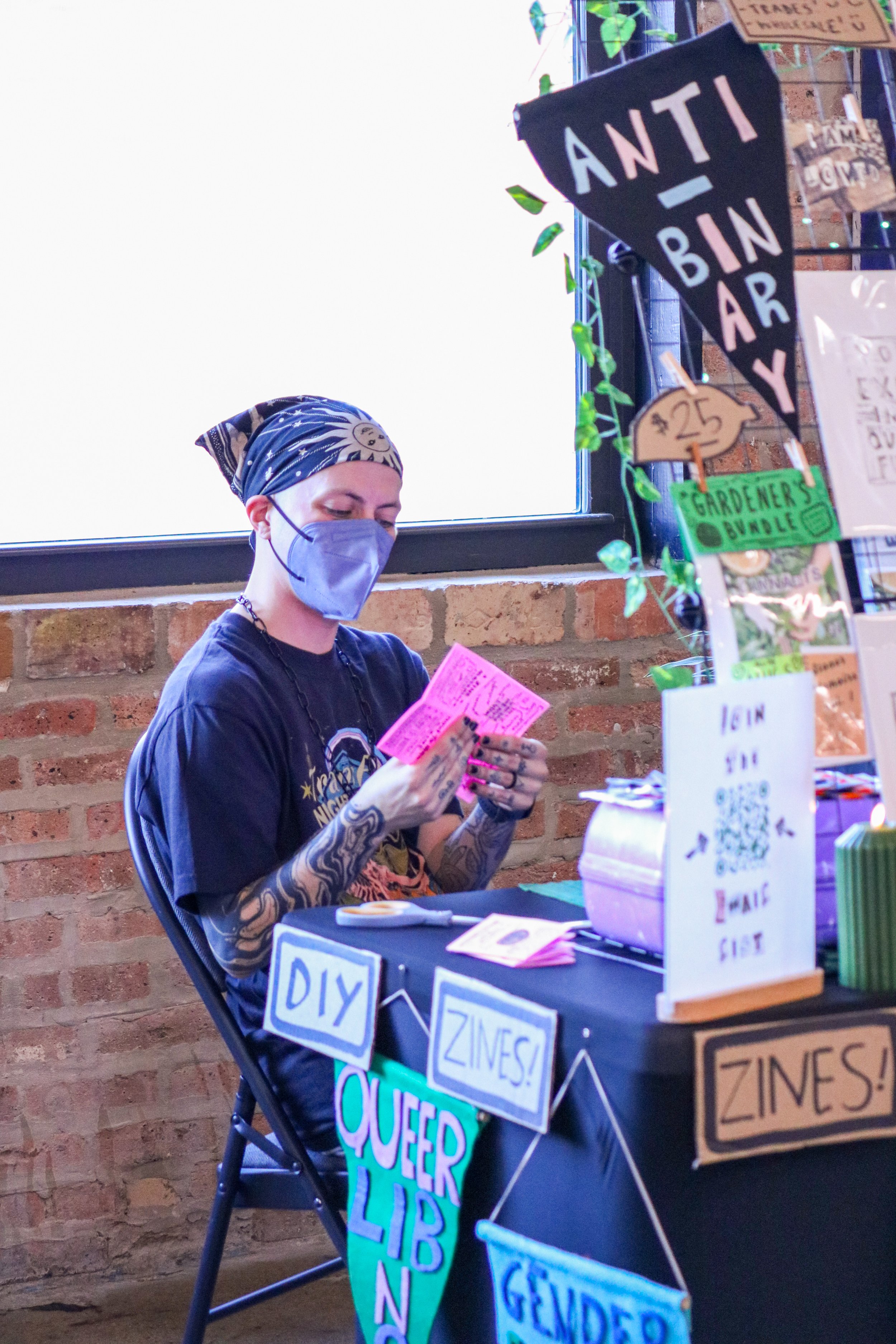 Wynter (side), wearing a mask, folding & packing zines at Witch Crafts Market, 2025, photo by @thatspoeticchi