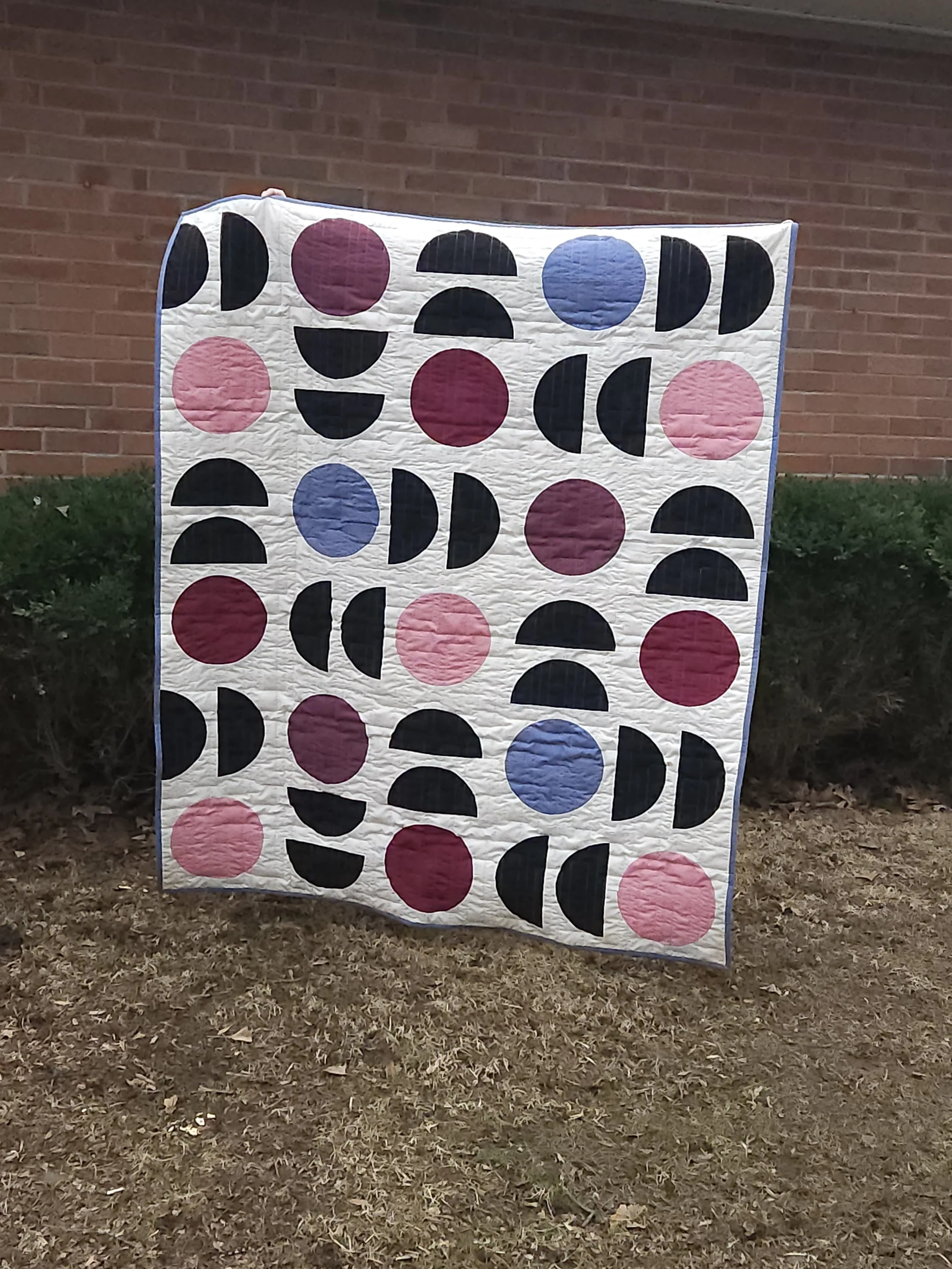 A quilt being held in front of shrubs and a brick wall. Quilt features circles and semi-circles of solid colored fabrics against a beige background.