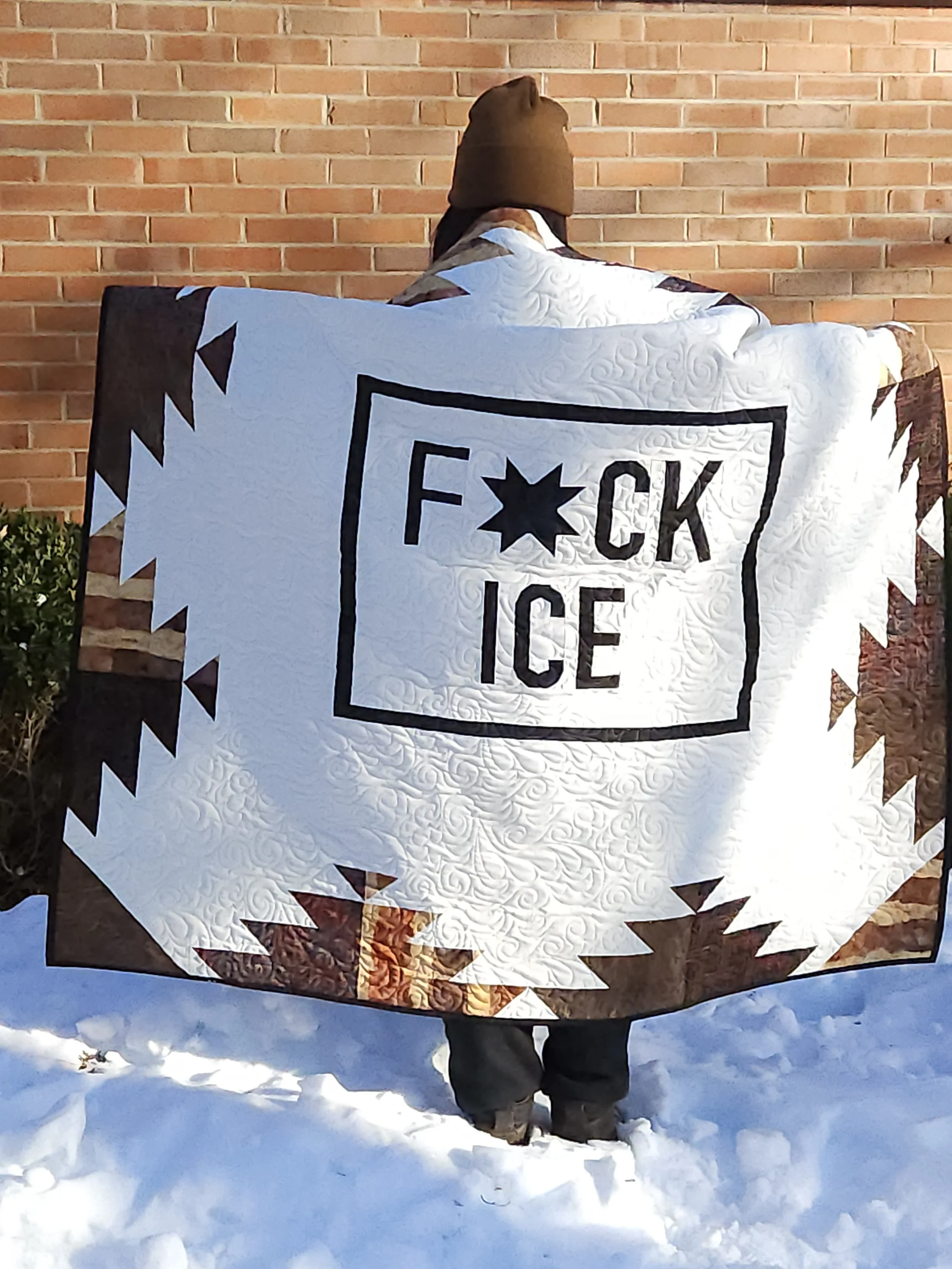 A woman with a hat standing in the snow, a lap sized quilt draped over her shoulders. The quilt says "F*ck ICE" in the center and is bordered by an Aztec-inspired geometric design.