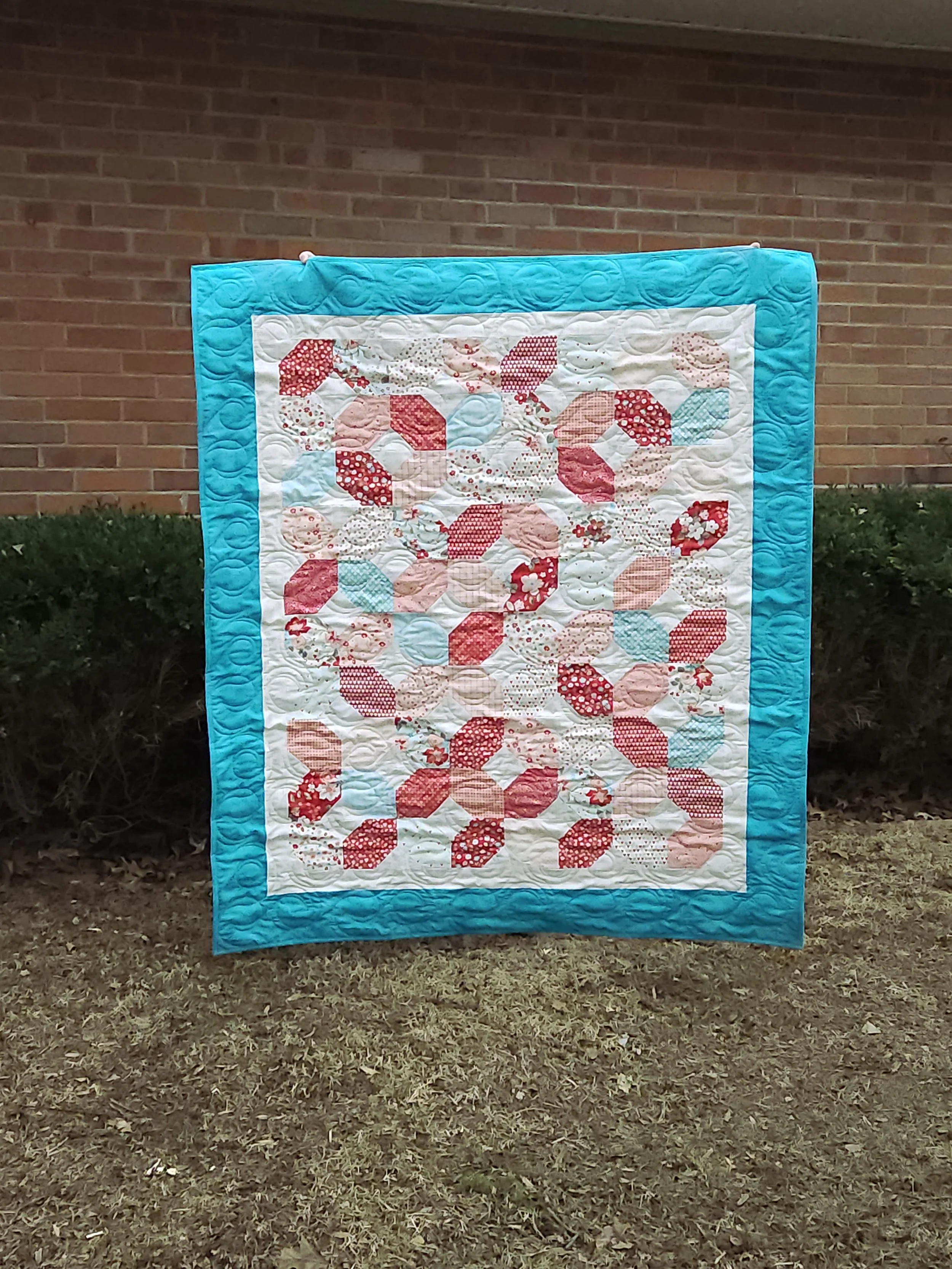 A quilt of red, pink & turquoise fabric creating a lattice effect over a white background with an outer, turquoise border. Quilt is being held in front of shrubs and a brick wall.