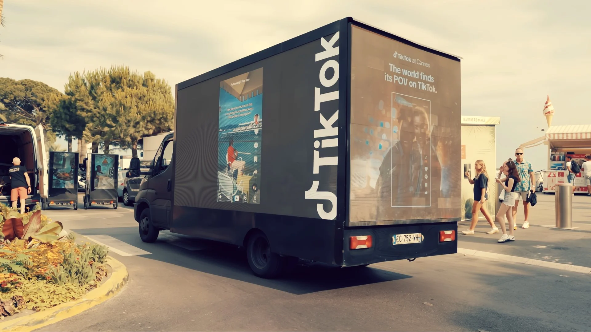 A black mobile billboard truck promoting TikTok at Cannes, displaying a large screen with TikTok content, and a group of people walking nearby in a parking lot with trees and a small ice cream stand in the background.