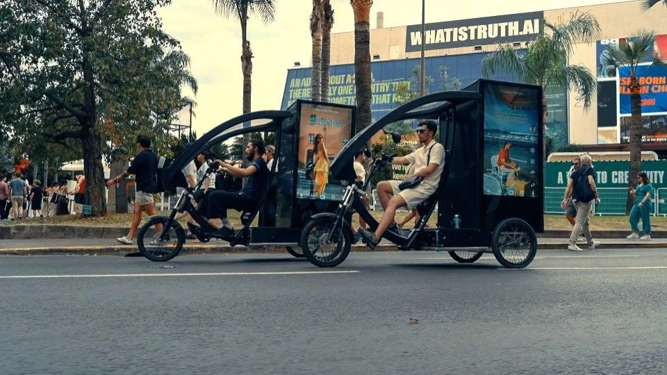 A tandem electric bike with two passengers, one man and one woman, riding down a city street. The bike has advertising on the back, and there are pedestrians on the sidewalk with trees and buildings in the background.