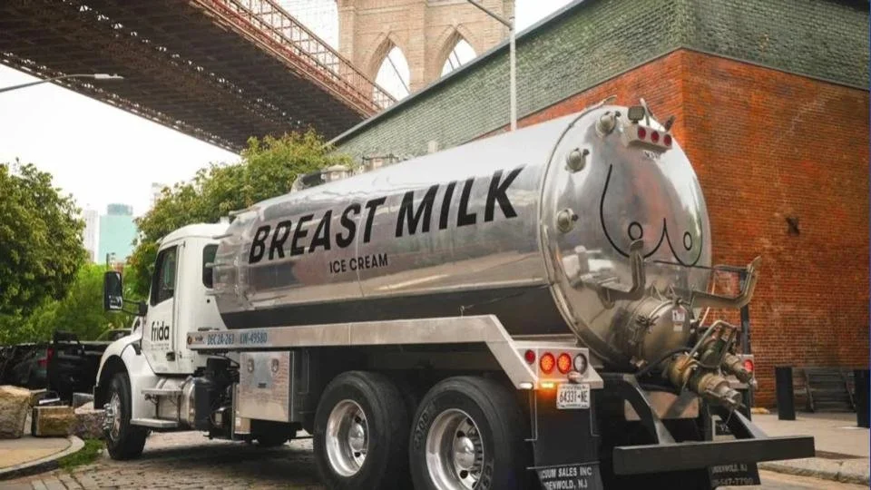 A milk tanker truck with the words 'Breast Milk Ice Cream' written on it, parked on a city street near some trees and a brick building.