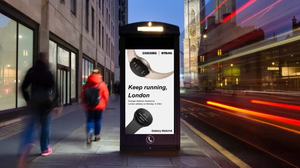 Street scene in London with blurred pedestrians walking past a digital billboard advertising a Samsung Galaxy Watch4, displaying text 'Keep running, London' and an image of the watch, near the Big Ben clock tower at dusk with streaks of light from pa