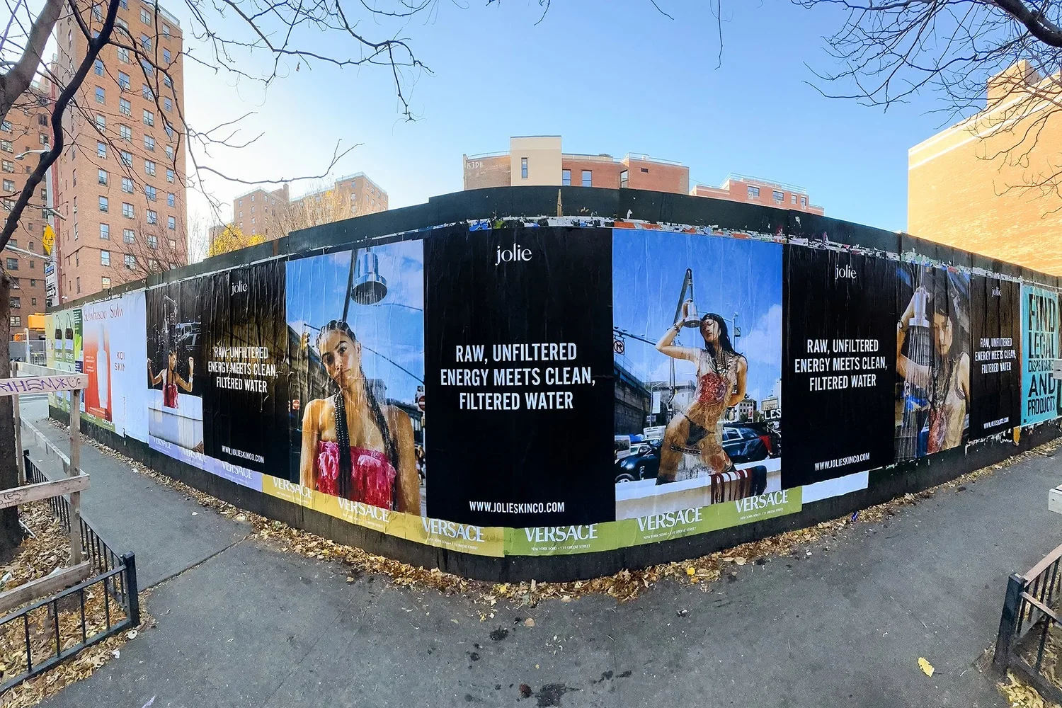 A city sidewalk with billboard advertisements for Jolie skincare and Versace, featuring models and text promoting filtered water and clean energy.