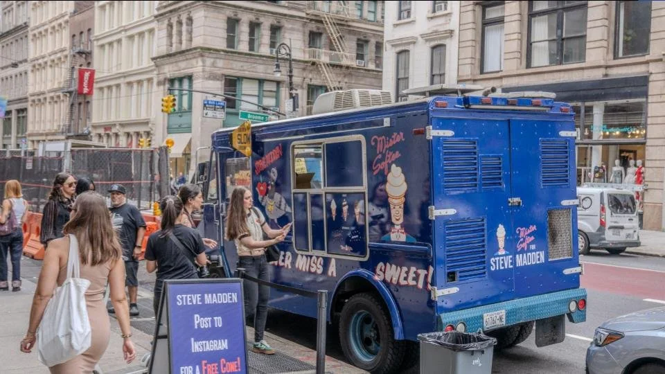 A blue ice cream truck with Steve Madden branding and ice cream graphics, parked on a city street. Several people are gathered near the truck, some ordering or talking, with tall buildings in the background.