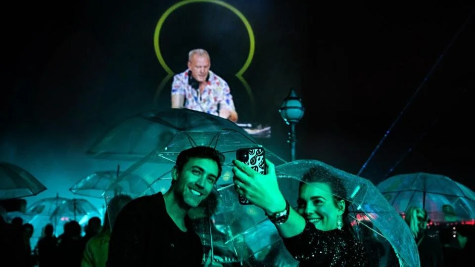 Two smiling women holding umbrellas taking a selfie at an event, with a DJ performing on stage in the background at night.