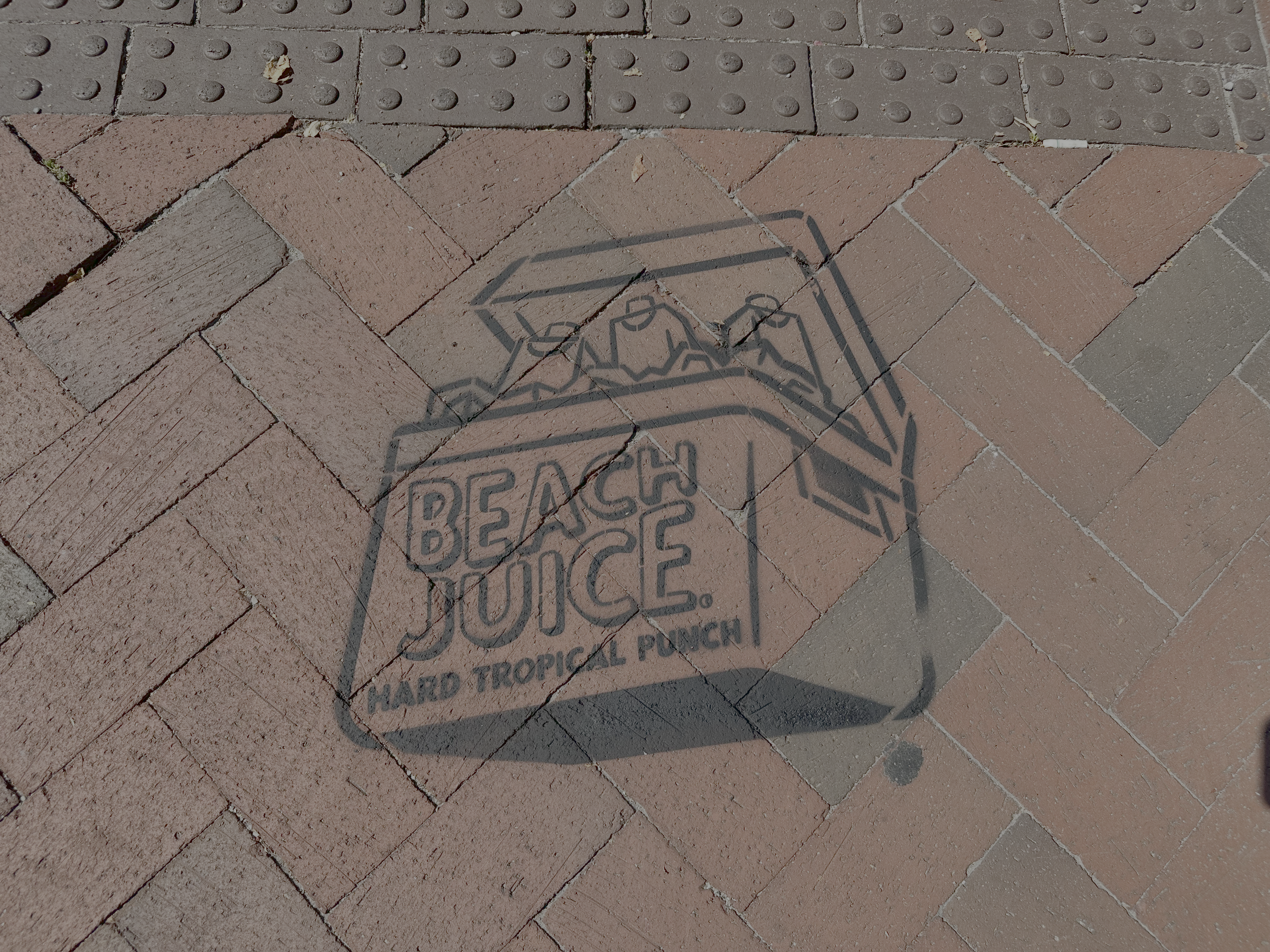 Shadow of a promotional sign on a brick pavement that reads 'Beach Juice Hard Tropical Punch' with a graphic of a box of juice.