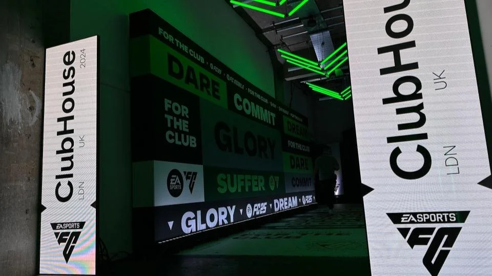 Indoor gaming event setup with large vertical screens displaying 'ClubHouse UK' and 'EA Sports FC,' green and black color theme, digital graphics, and people walking nearby.