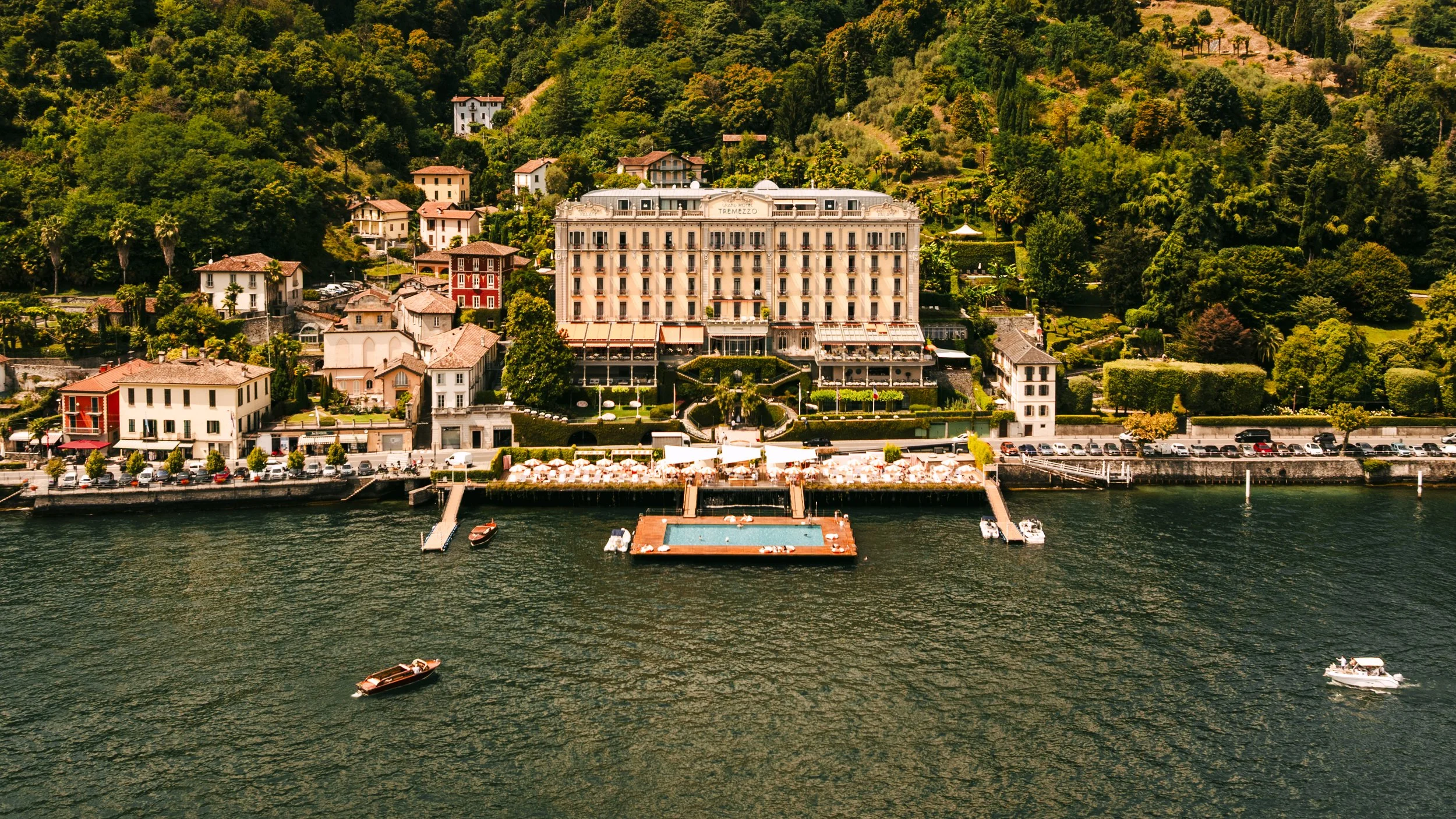 Large hotel building with outdoor pool, situated along a lake with boats, and surrounded by lush green hills and residential houses.