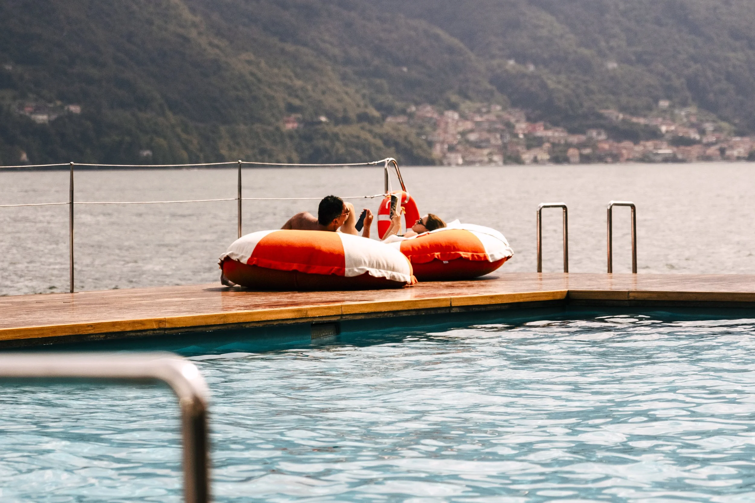 Two people relaxing on bean bags by the poolside, with a lake and hills in the background.