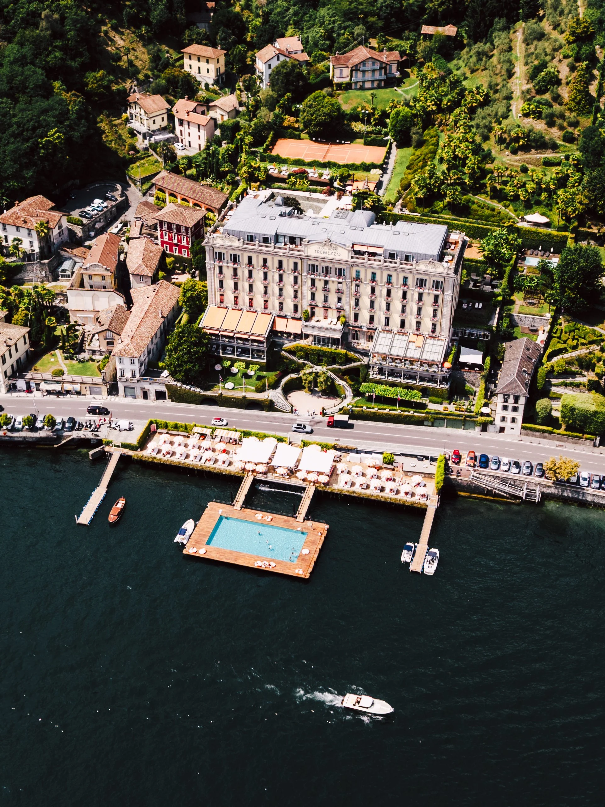 Aerial view of a lakeside hotel with a swimming pool and boats docked at the marina, surrounded by green trees and residential buildings.