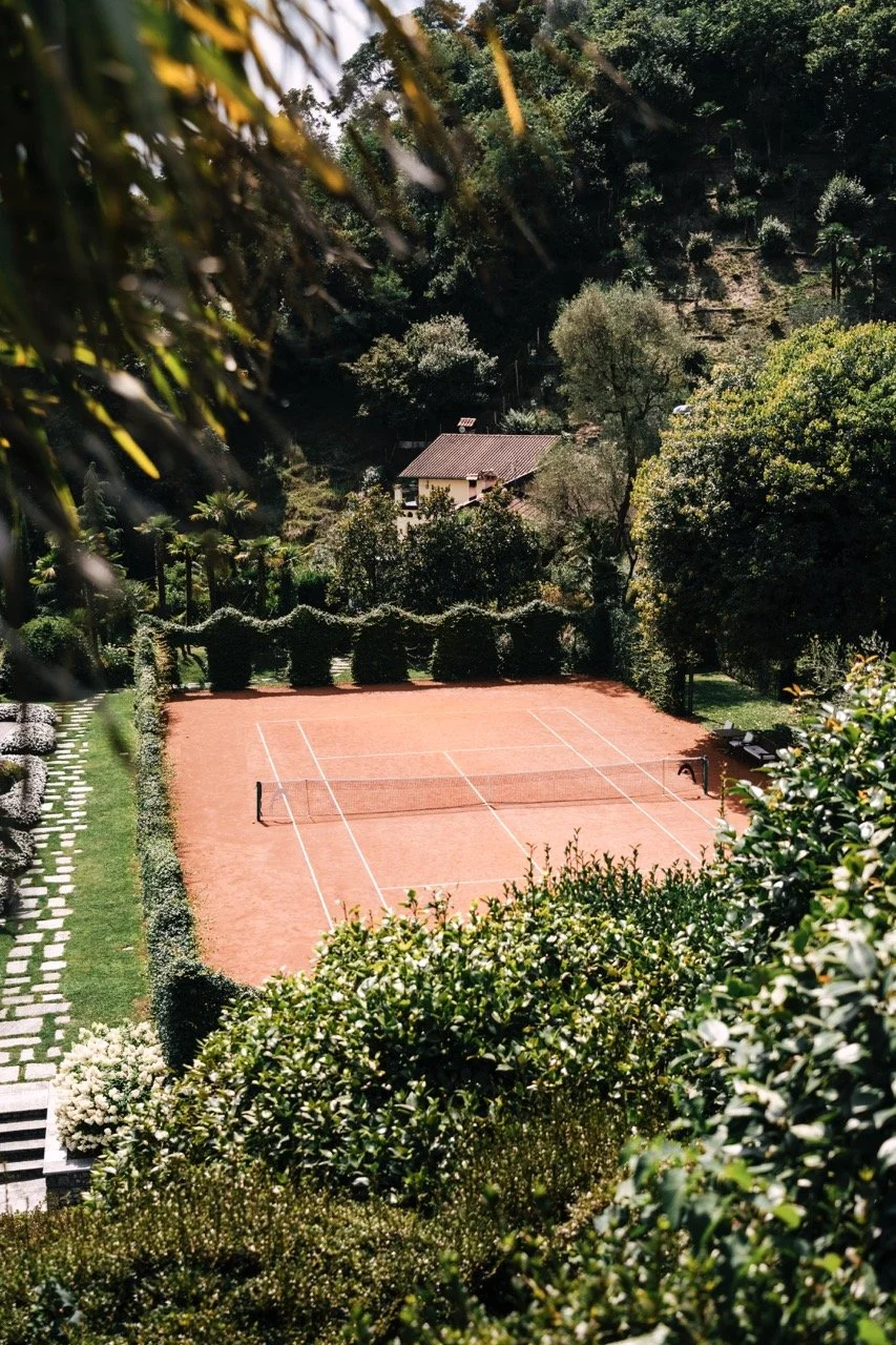Red clay tennis court surrounded by lush greenery and trees, with a small house in the background, under a clear sky.