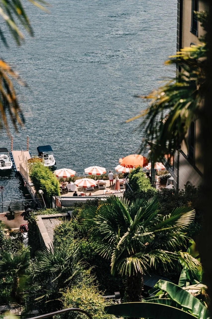 View of a waterfront patio area with orange and white umbrellas, people sitting and walking, boats docked at a pier, and lush green tropical plants in the foreground.