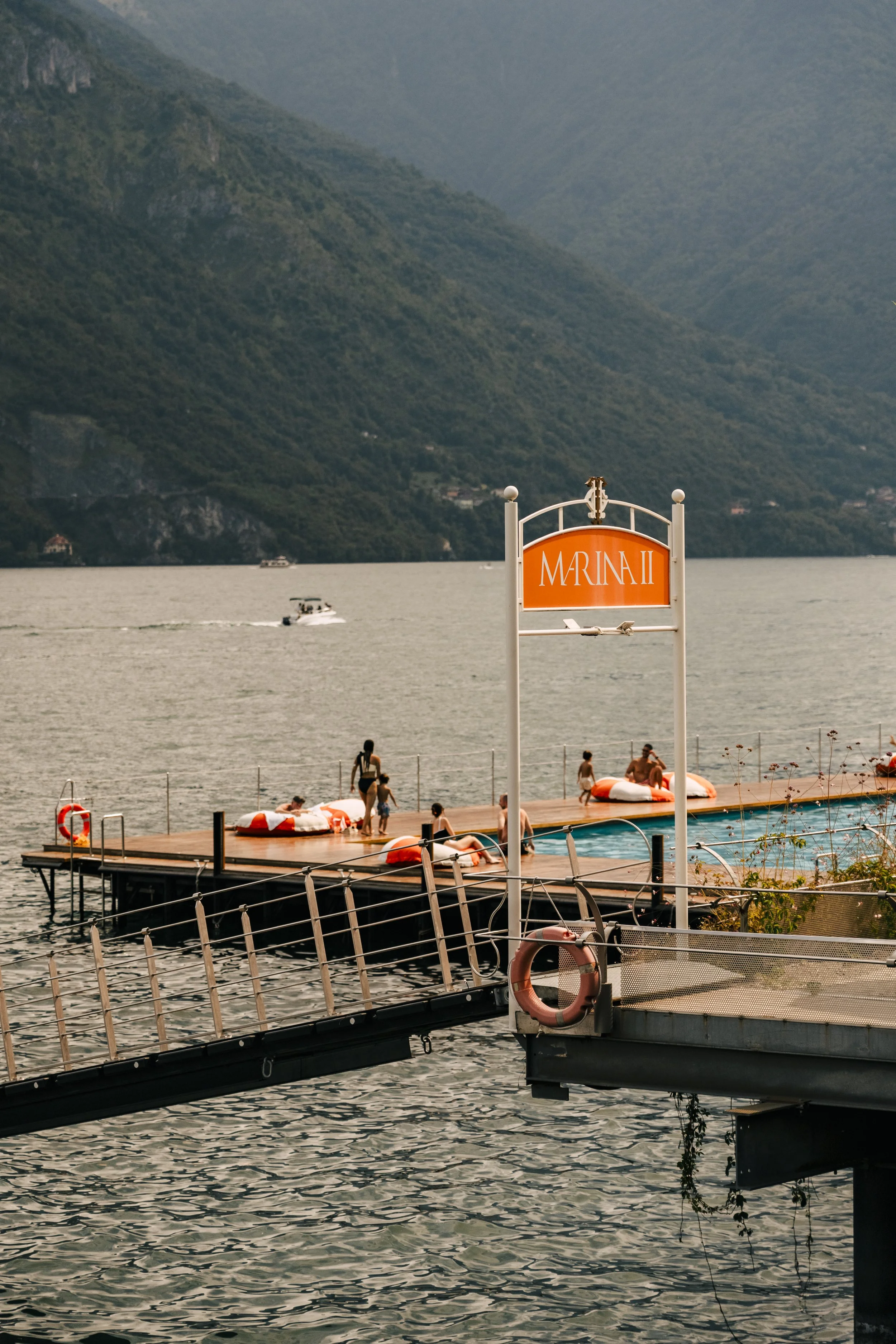 People relaxing and swimming at a lakeside dock with a sign that reads 'MARINA II,' surrounded by mountains, during overcast weather.