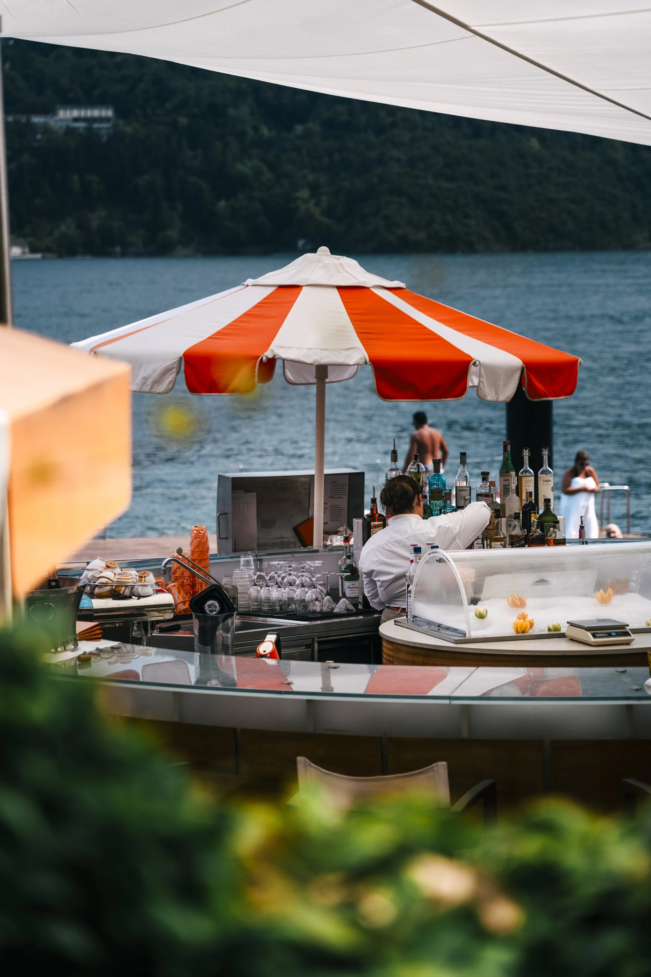A lakeside bar with a red and white striped umbrella, bottles of alcohol, a bartender, and people near the water.