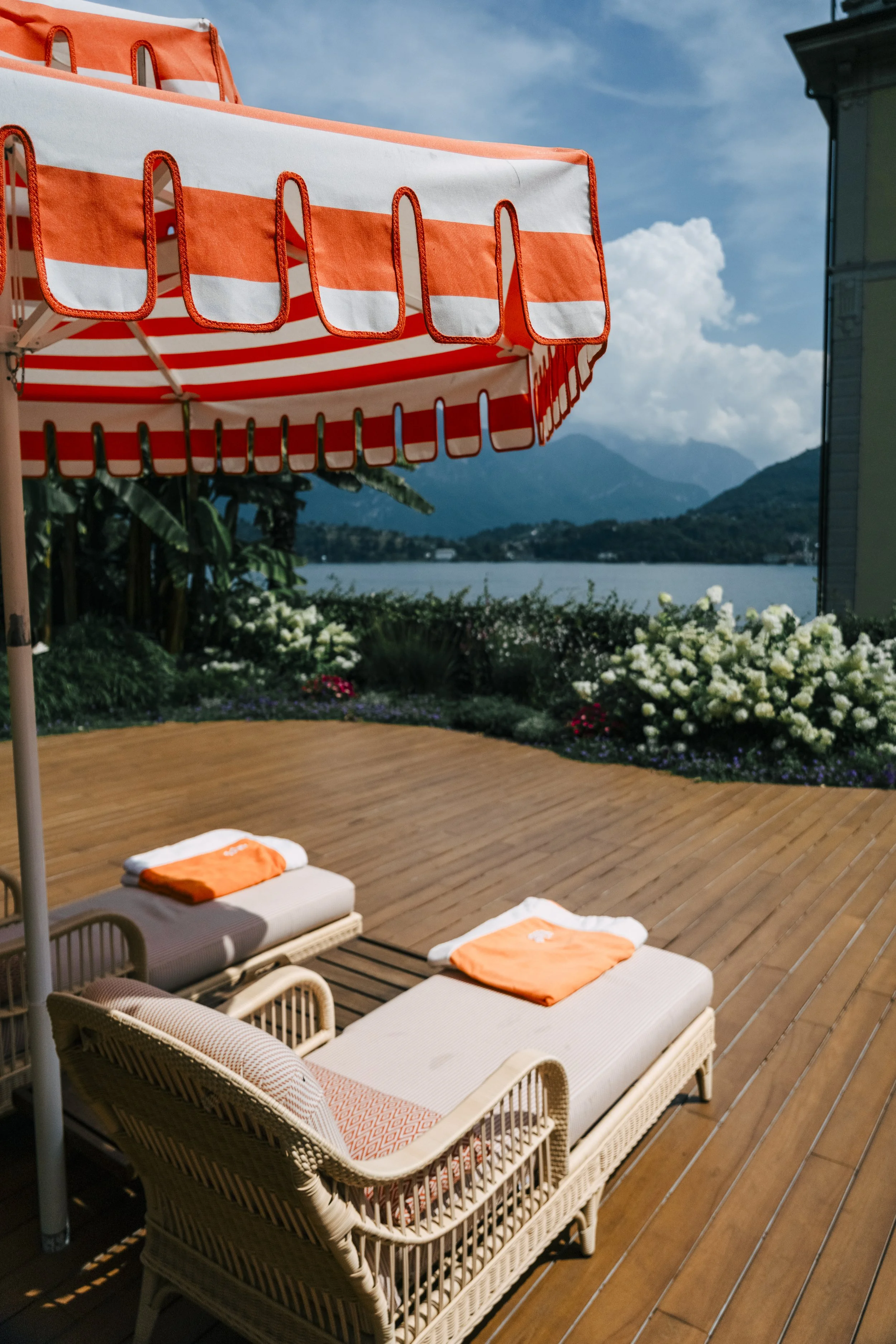 Outdoor deck with two lounge chairs, orange towels, and a large striped orange and white umbrella. Flowers and vegetation near a body of water with mountains in the background.