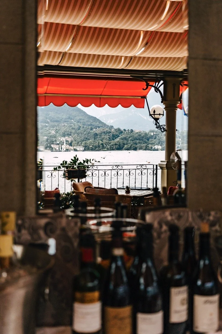 View through a restaurant window showing an outdoor patio with chairs and tables, overlooking a lake with mountains in the background, and a red awning overhead.