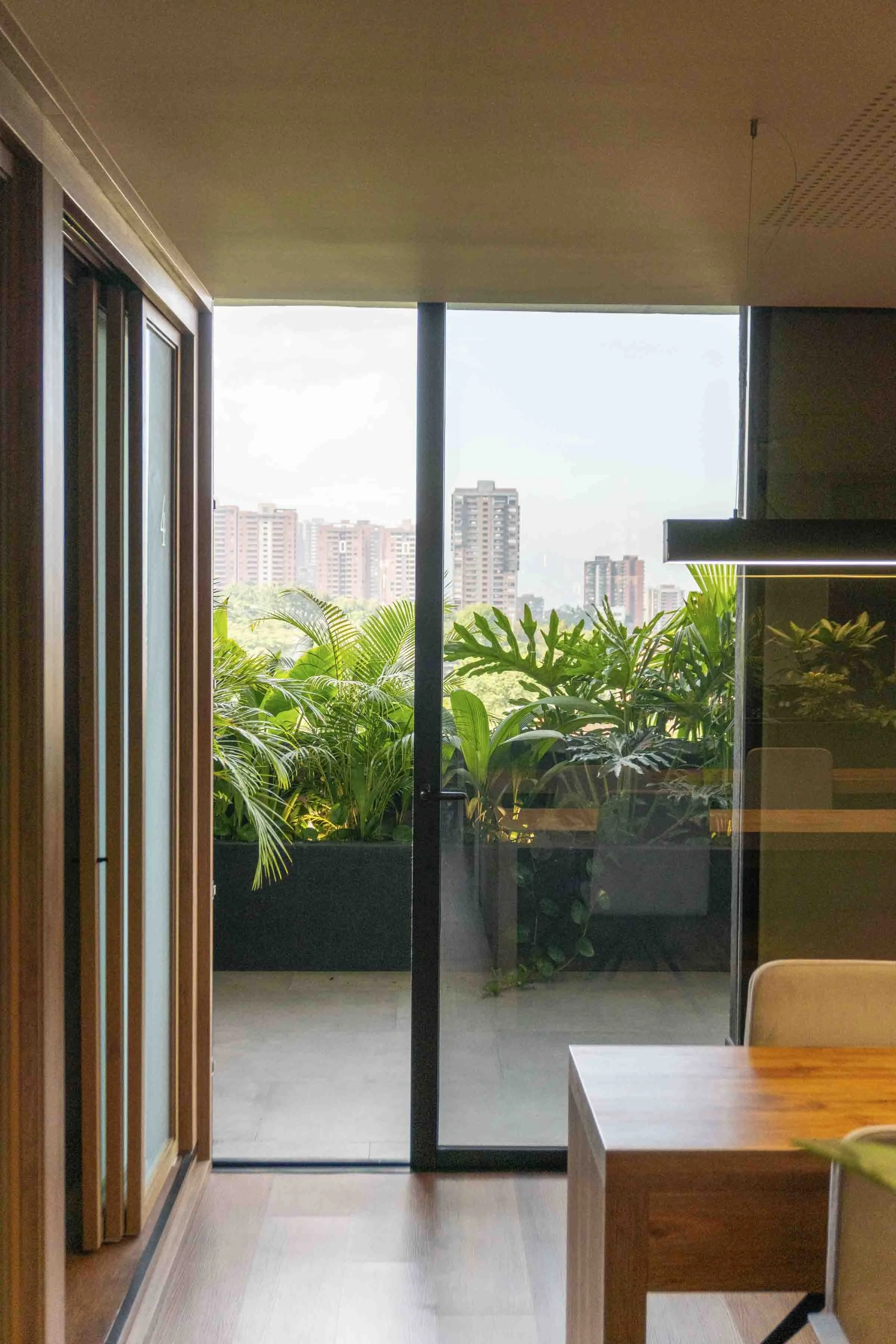 Office interior with wooden table and glass door leading to a balcony with potted plants and city skyline in the background.