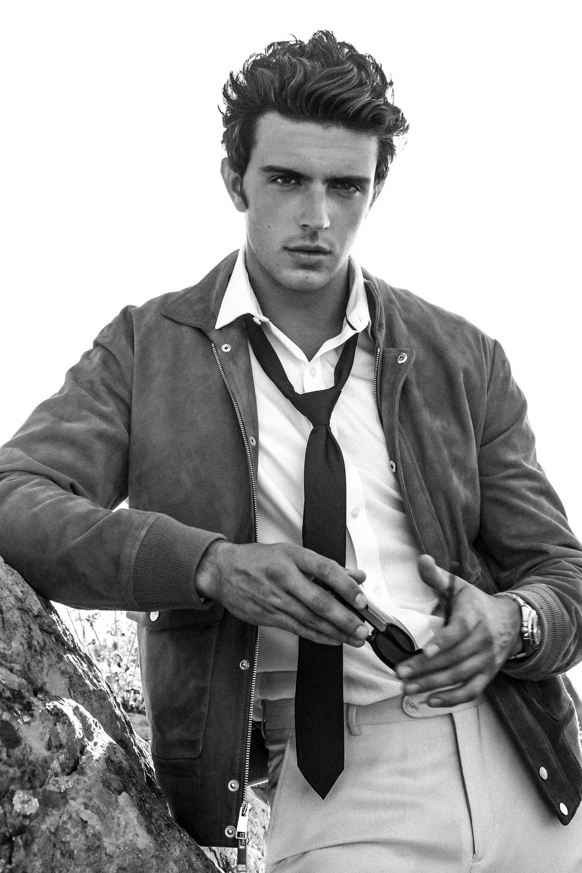 Black and white photo of a young man with styled hair, wearing a white shirt, black tie, and a jacket. He is holding sunglasses and leaning against a tree.