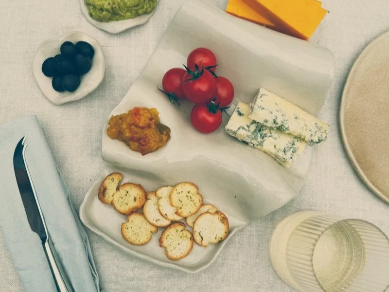 A cheese board with cherry tomatoes, blue cheese, crackers, small bowl of blueberries, and a wedge of cheddar cheese on a table with a beige plate, drinking glass, and knife on a napkin.