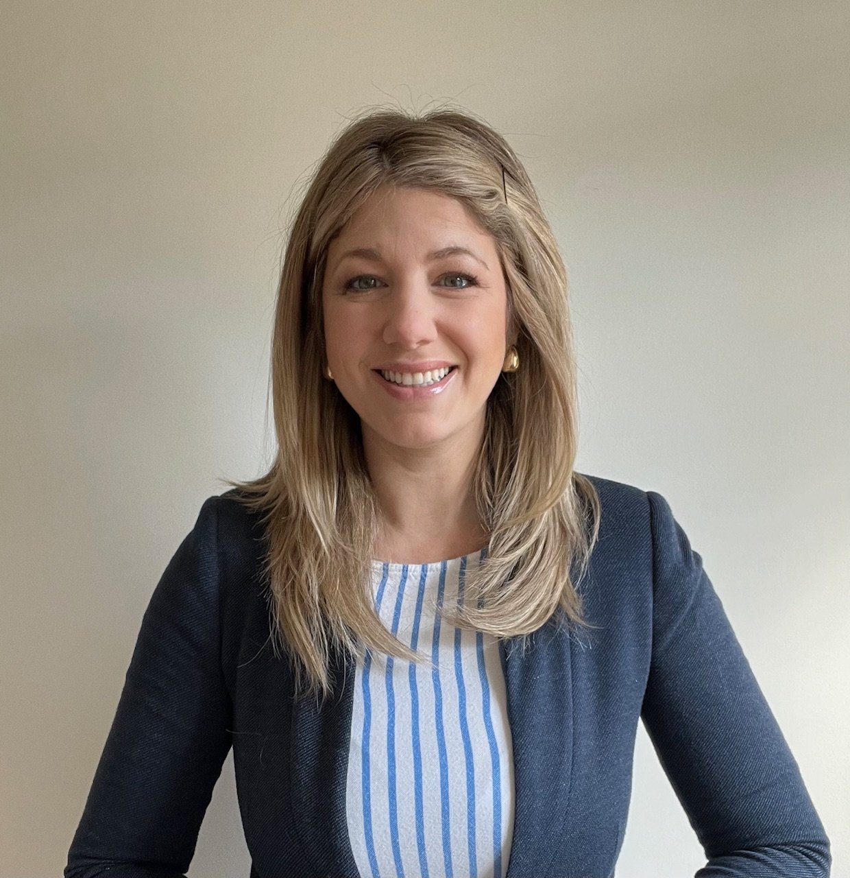 A woman with shoulder-length blonde hair, wearing a navy blazer and a light blue and white striped blouse, smiling at the camera.