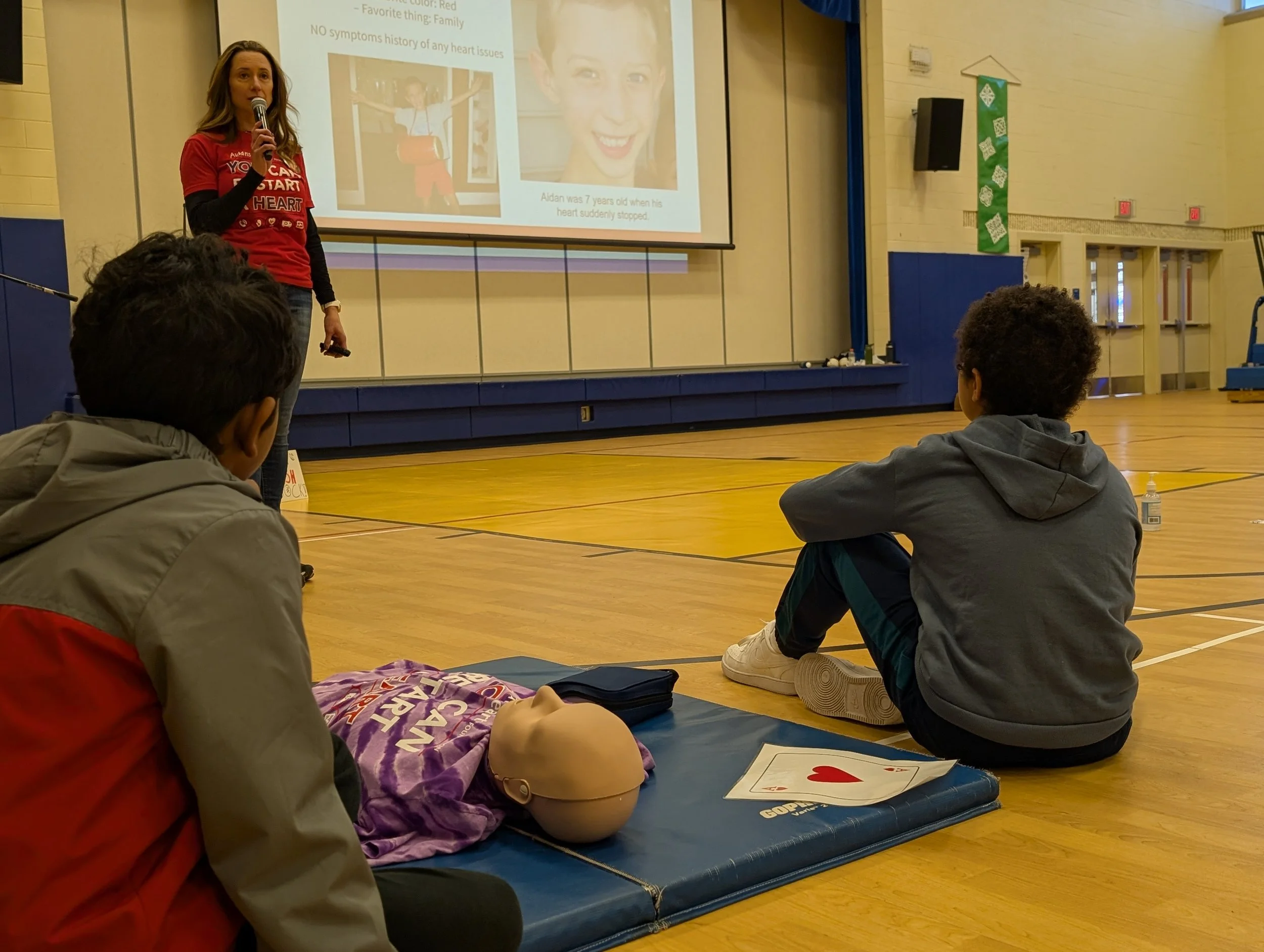 A woman giving a CPR training presentation in a gymnasium with two young boys seated on the floor. The boys are watching a CPR mannequin and an oversized playing card with a red heart. A large screen behind the woman displays a slide with images and text about heart health.