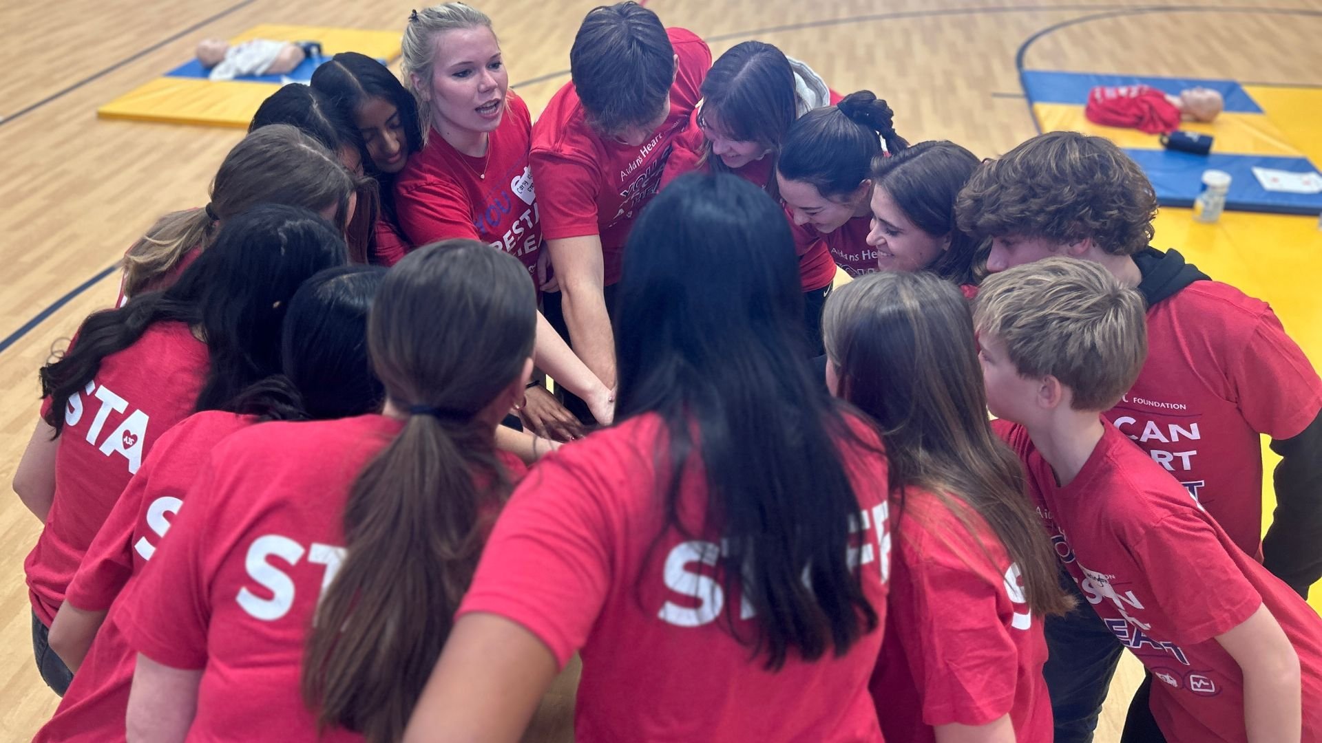 Group of people wearing red shirts huddled together in the center of a gymnasium, participating in a team activity.