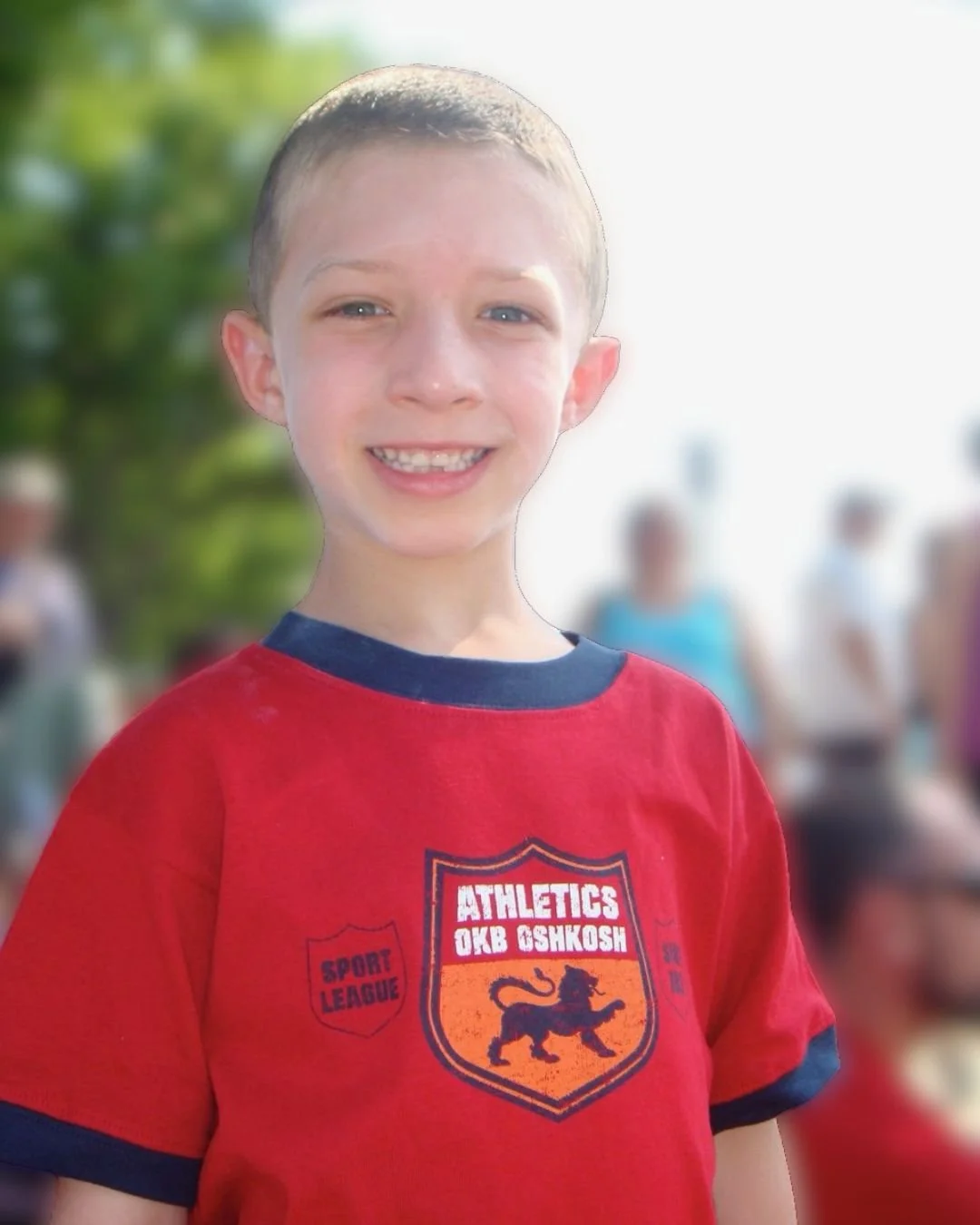 Young boy smiling outdoors, wearing a red T-shirt with a black collar and a sports logo.