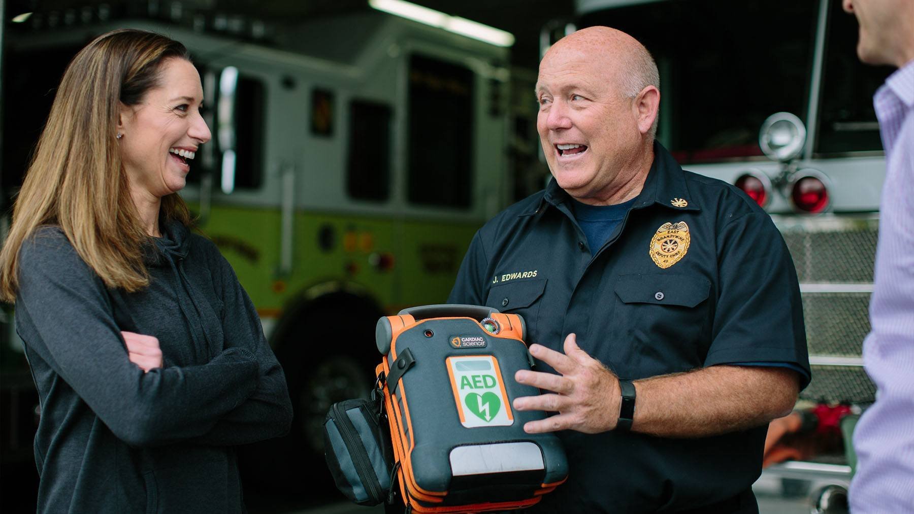 A man in a fire department uniform explaining a portable AED device to a woman with crossed arms, all standing outdoors in front of a fire truck.
