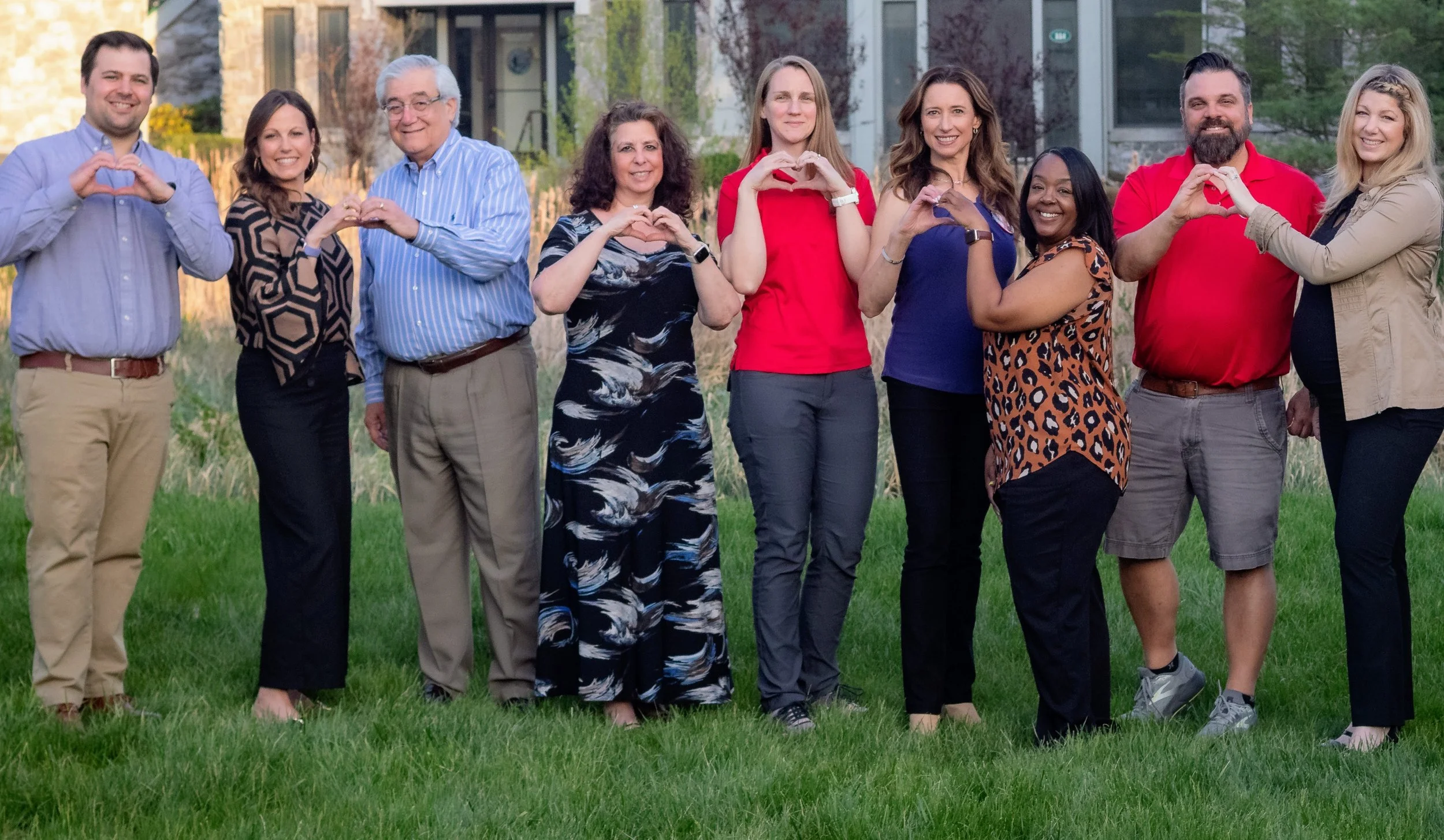 Group of nine diverse people standing on grass outdoors, making heart shapes with their hands.