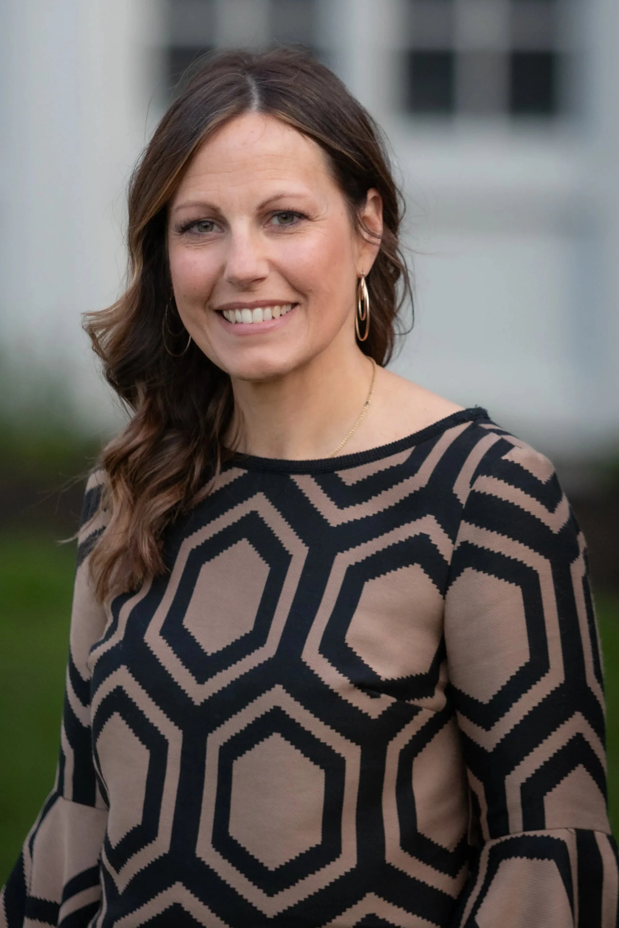 A woman with brown hair wearing a black and beige geometric patterned top, gold jewelry, and earrings, smiling outdoors in front of a building.