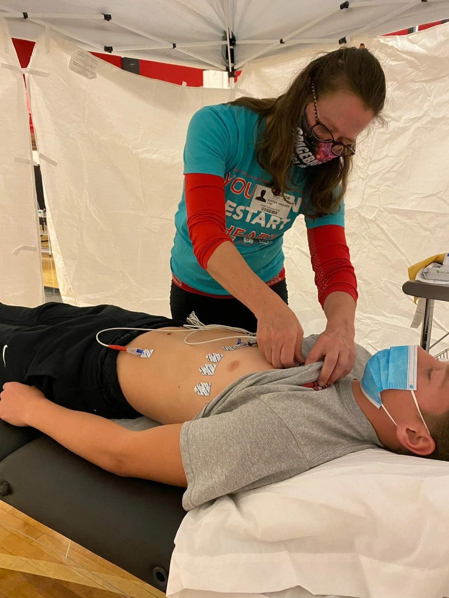 A woman giving cardiac care to a young man lying on a medical bed. The young man is wearing a face mask, and the woman is attaching electrodes to his chest for a medical procedure.