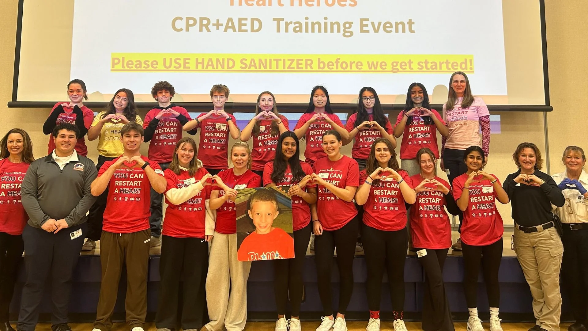 Group of young people and adults on stage during CPR/AED training event, some wearing red shirts with message 'You Can Restart A Heart', holding a poster of a smiling boy, with a large screen behind them displaying event information and a reminder to use hand sanitizer.