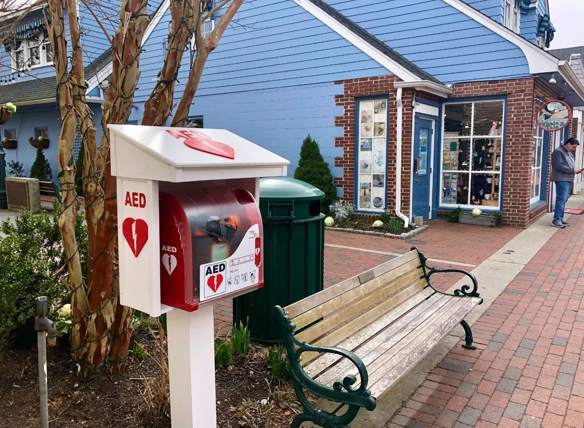 Medical AED emergency kit box on a sidewalk next to a park bench and a tree, with a blue building and a person in the background.