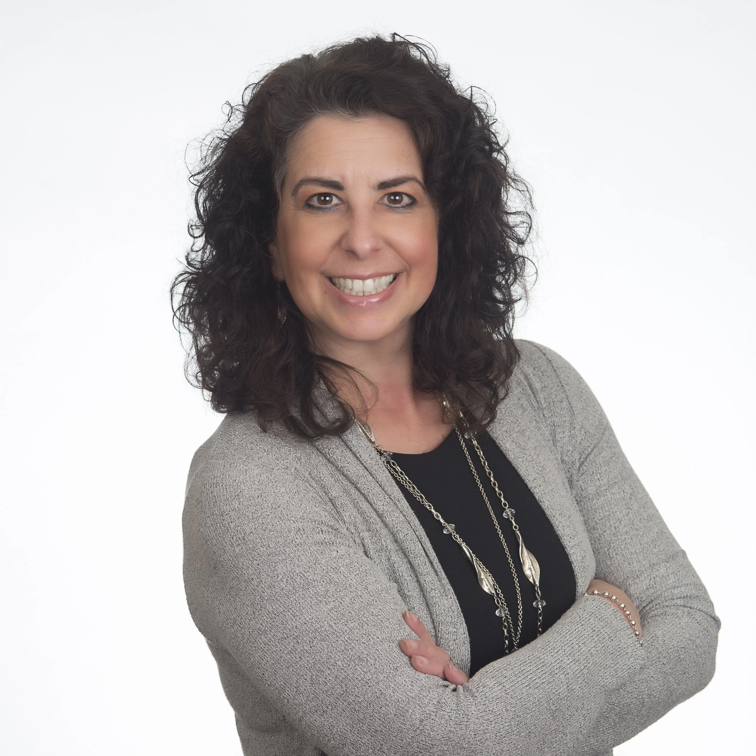 A smiling woman with dark curly hair, wearing a gray blazer over a black top, with layered necklaces and crossed arms, posing against a white background.