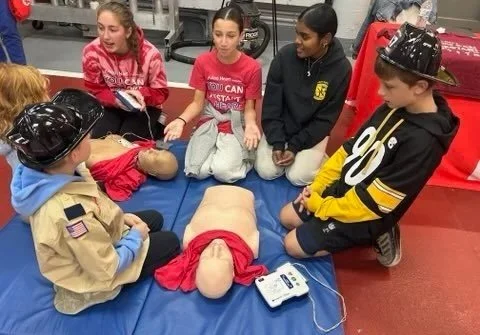 Children sitting around a CPR training mannequin on a blue mat, participating in a first aid demonstration in a school gym.