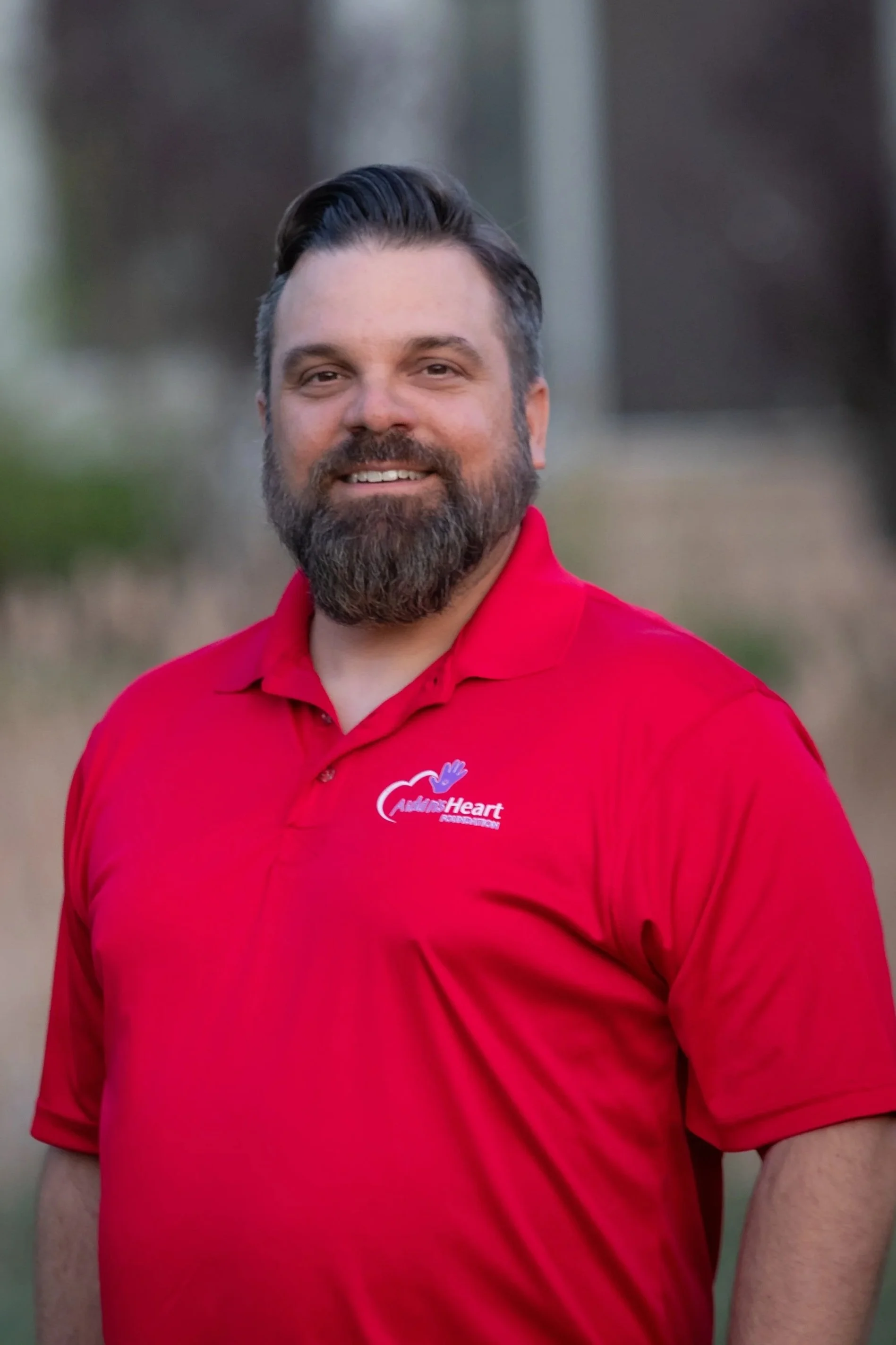 A man with a beard and dark hair, wearing a red polo shirt with a logos on chest, smiling outdoors with a blurred background.