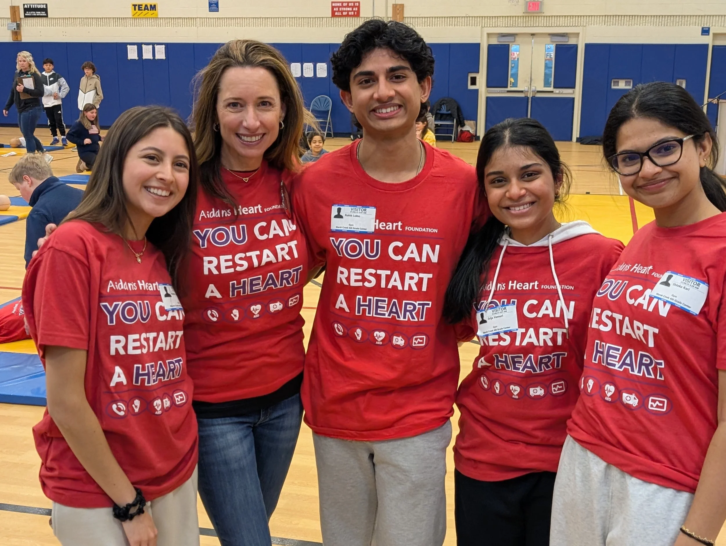 Group of five people, four young women and one young man, standing together indoors in a gym. They are all wearing red T-shirts with the slogan 'You Can Restart a Heart' and logos related to a heart foundation. Two of the women and the young man have visitor badges. They are smiling and posing for the photo.