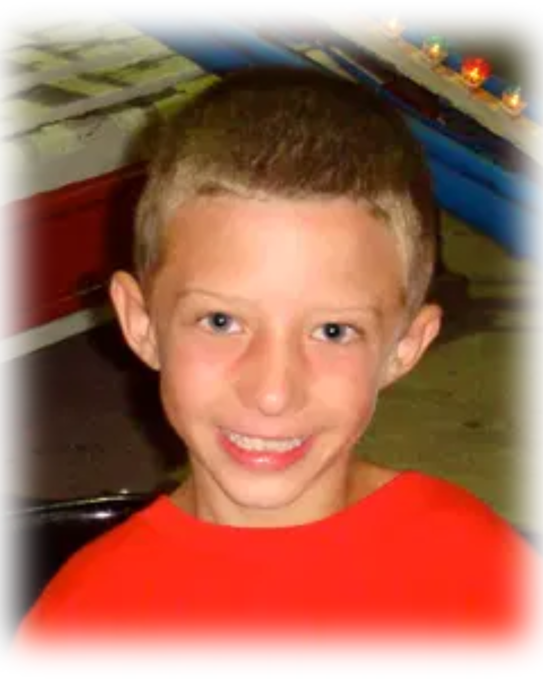A smiling young boy with short brown hair and blue eyes, wearing a red shirt, sitting in a classroom with colorful bins and supplies in the background.