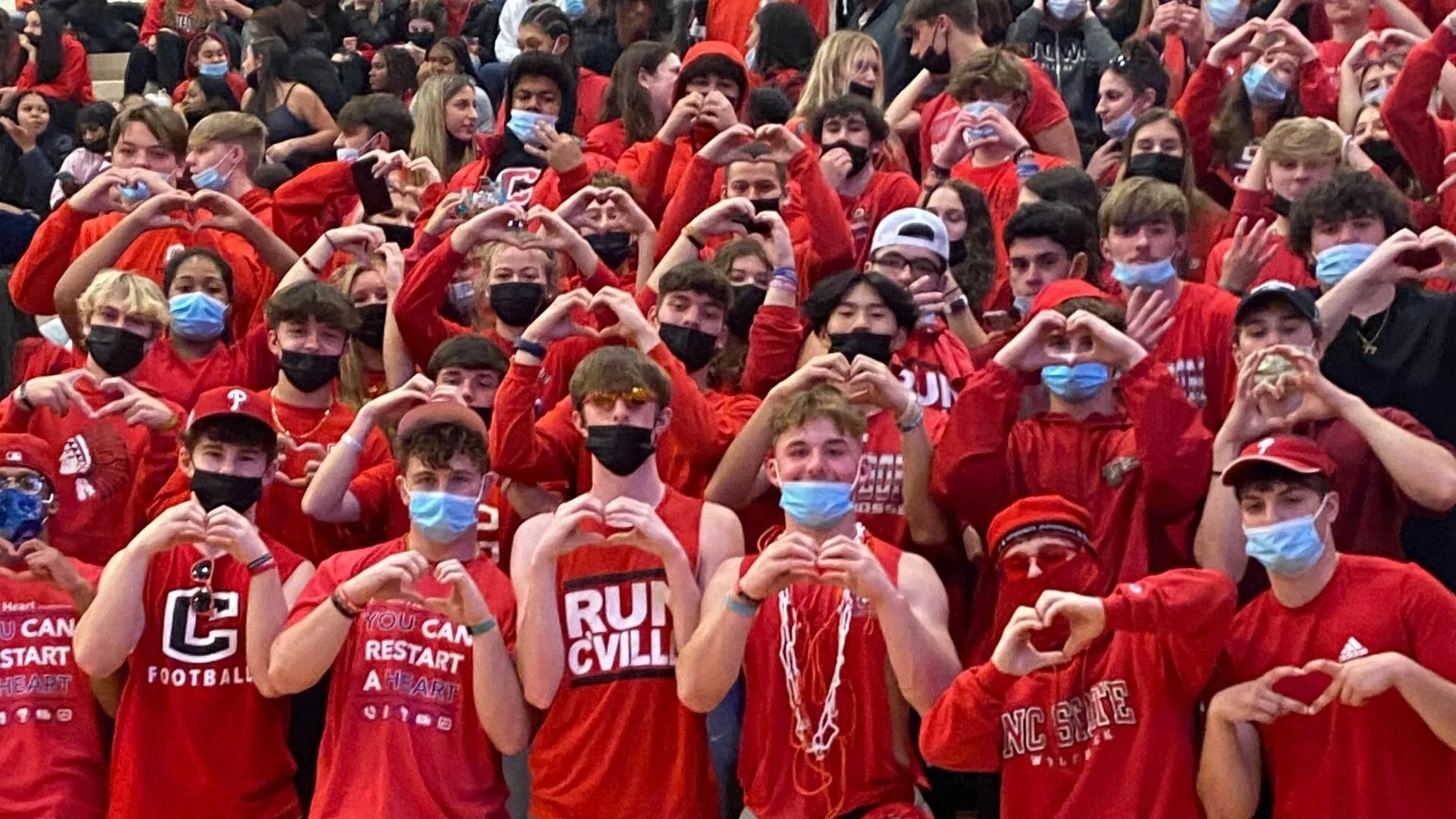 A large group of young people wearing red shirts and face masks, many making heart shapes with their hands, gathered at an indoor event.