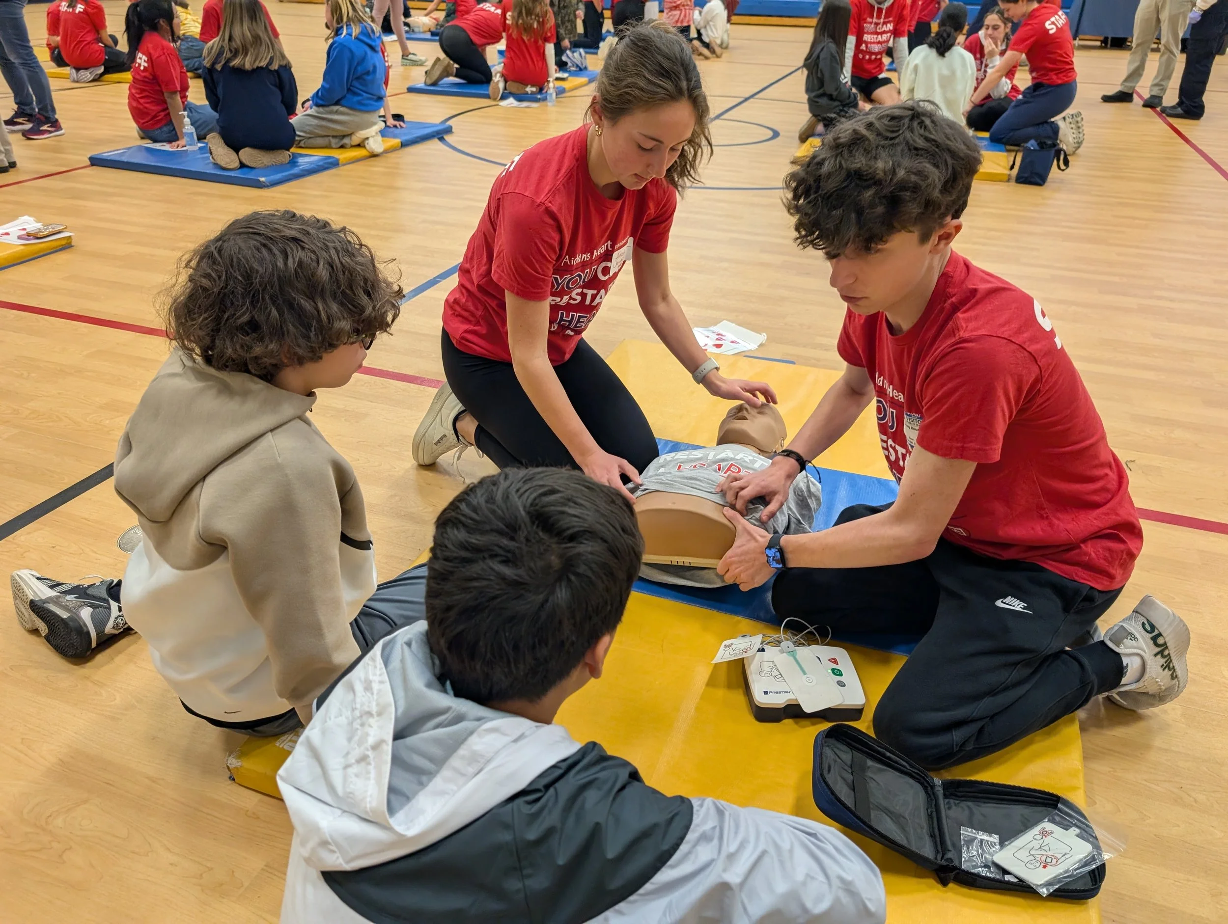 Group of young people participating in a CPR training session in a gymnasium, with two instructors demonstrating on a CPR manikin.
