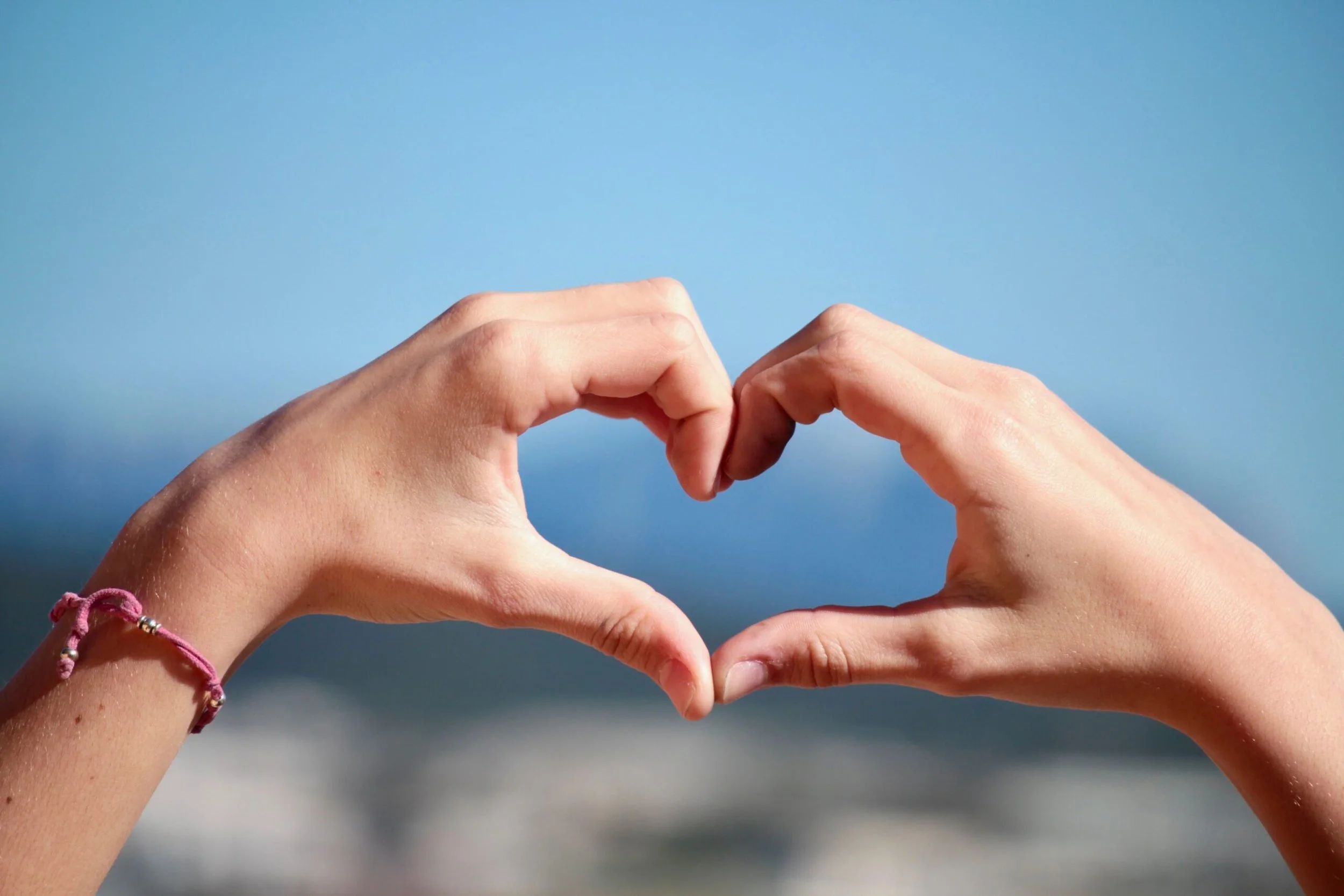 Hands forming a heart shape against a blue sky and ocean background.