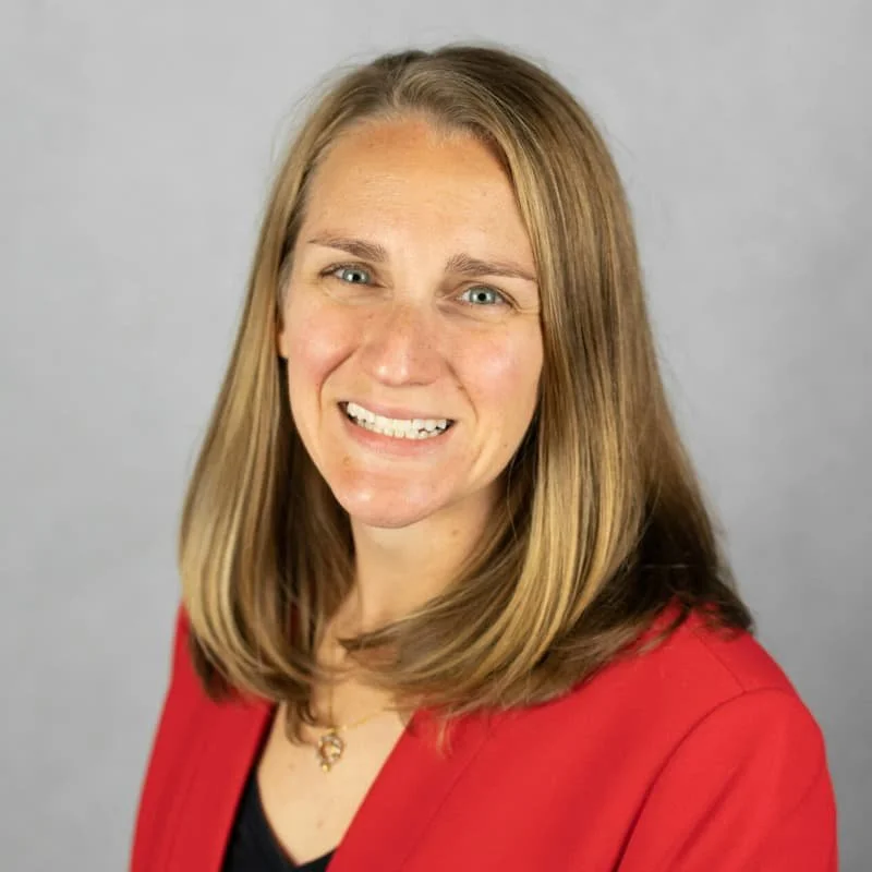 A woman with long blonde hair, smiling, wearing a red blazer and a black top, against a gray background.